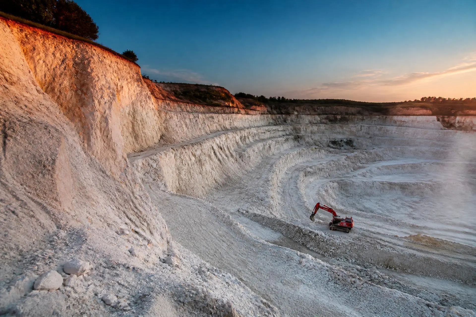 Open-pit kaolin clay quarry with terraced white clay walls and a red excavator at dusk